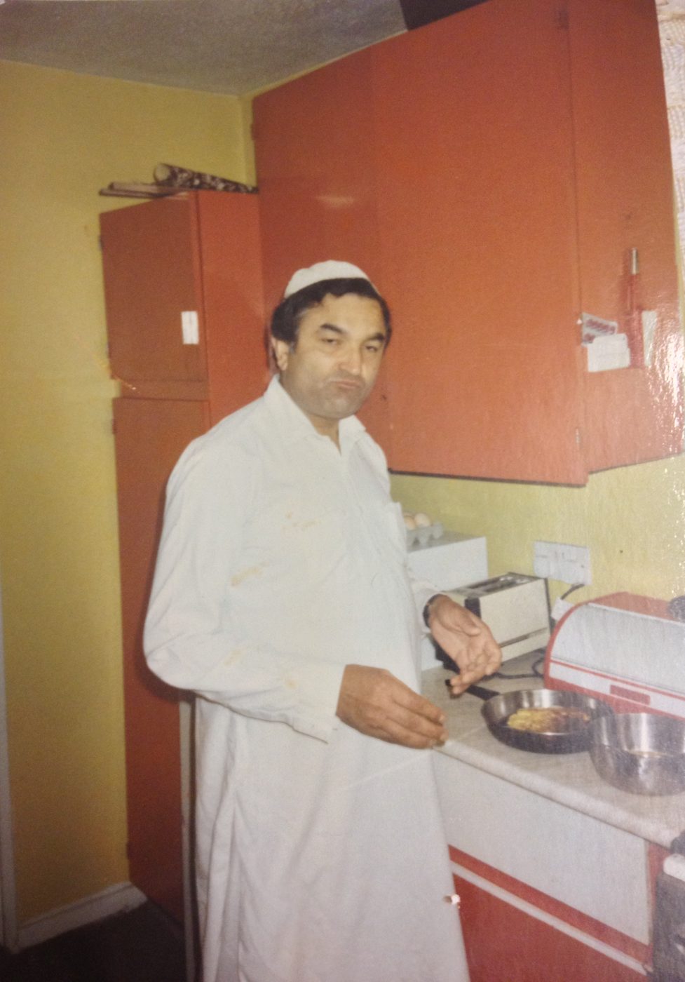 Mukhtar Ahmed in a white kameez and prayer cap, standing in the kitchen at home
